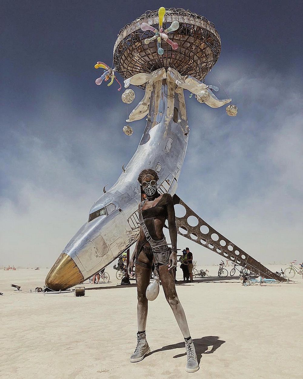Участница в черепной маске, покрытая playa dust, рядом с арт-объектом